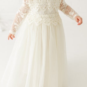 May include: A floor-length cream-colored dress featuring long sheer lace sleeves, a high neckline with scalloped lace trim, and a bodice adorned with intricate floral lace embroidery. The skirt is made of layered tulle fabric.