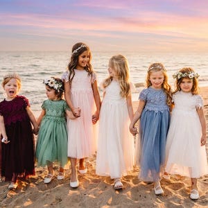 May include: Five children standing on a sandy beach wearing floor-length dresses with lace bodices and tulle skirts in shades of burgundy, sage green, light pink, white, and dusty blue, each accessorized with a floral crown on their head.