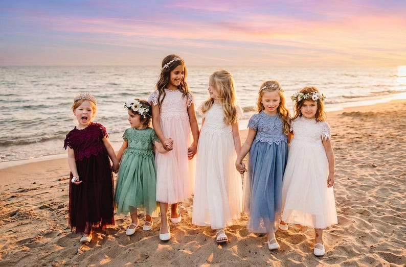 May include: Five children standing on a sandy beach wearing floor-length dresses with lace bodices and tulle skirts in shades of burgundy, sage green, light pink, white, and dusty blue, each accessorized with a floral crown on their head.