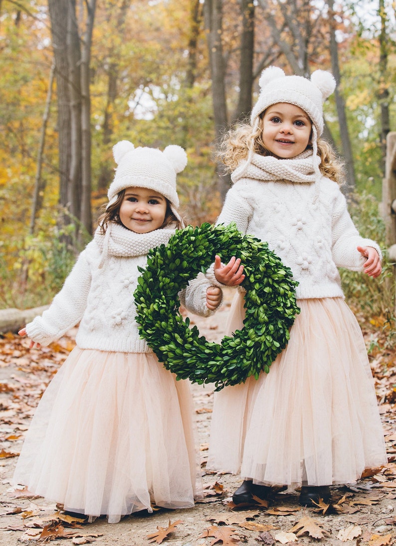 May include: Two young girls wearing white knit sweaters and matching tulle skirts with white pom-pom hats. The girls are holding a green boxwood wreath.