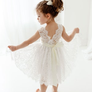 Pode incluir: Uma menina pequena vestindo um vestido branco de renda com uma faixa de cetim. O vestido tem costas baixas e uma bainha festonada. A menina est&aacute; descal&ccedil;a e tem o cabelo preso em um coque com um la&ccedil;o branco.