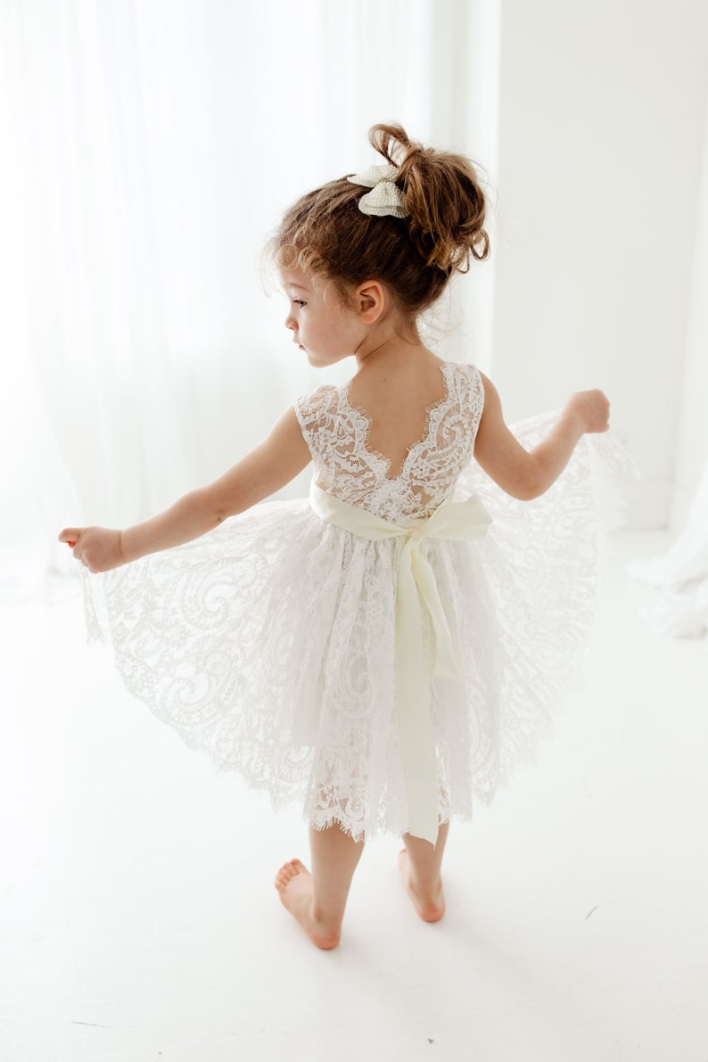 Pode incluir: Uma menina pequena vestindo um vestido branco de renda com uma faixa de cetim. O vestido tem costas baixas e uma bainha festonada. A menina est&aacute; descal&ccedil;a e tem o cabelo preso em um coque com um la&ccedil;o branco.
