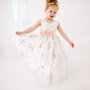 Puede incluir: Una ni&ntilde;a peque&ntilde;a con un vestido de tul blanco con estampado floral y una faja de sat&eacute;n melocot&oacute;n. El vestido tiene un corpi&ntilde;o de encaje y una falda completa. La ni&ntilde;a lleva una diadema dorada con un adorno floral.