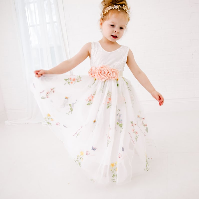 Puede incluir: Una ni&ntilde;a peque&ntilde;a con un vestido de tul blanco con estampado floral y una faja de sat&eacute;n melocot&oacute;n. El vestido tiene un corpi&ntilde;o de encaje y una falda completa. La ni&ntilde;a lleva una diadema dorada con un adorno floral.