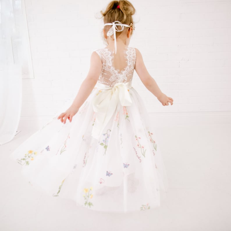 Puede incluir: Un vestido de tul blanco con un corpi&ntilde;o de encaje y estampado floral. El vestido tiene una cinta de sat&eacute;n atada alrededor de la cintura y una diadema blanca con una flor.