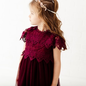 May include: A young girl wearing a burgundy tulle dress with a lace bodice and a floral crown.