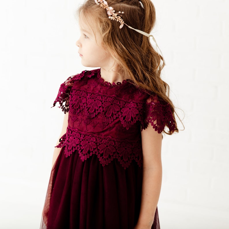 May include: A young girl wearing a burgundy tulle dress with a lace bodice and a floral crown.