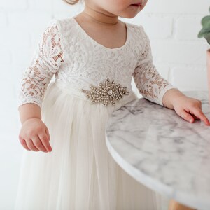 Pu&ograve; includere: Un abito bianco di tulle con maniche di pizzo e una cintura gioiello. L'abito &egrave; indossato da una bambina che sta in piedi vicino a un tavolo.