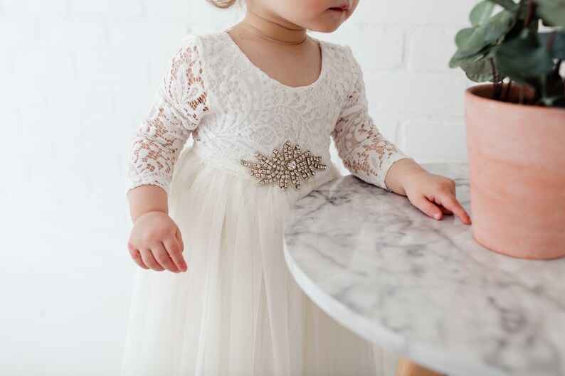 Pu&ograve; includere: Un abito bianco di tulle con maniche di pizzo e una cintura gioiello. L'abito &egrave; indossato da una bambina che sta in piedi vicino a un tavolo.