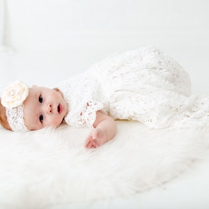 K&ouml;nnte beinhalten: Ein Baby in einem wei&szlig;en Spitzenkleid und einem Stirnband mit einer wei&szlig;en Blume, das auf einer wei&szlig;en, flauschigen Oberfl&auml;che liegt.
