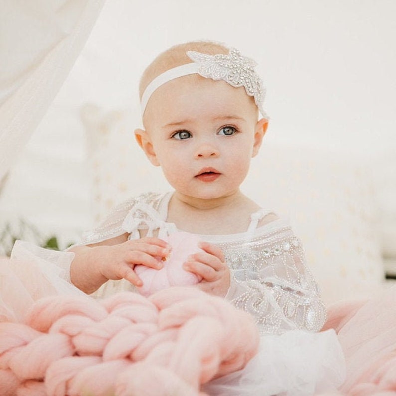 May include: A baby girl wearing a white dress with a beaded overlay and a white headband with a sparkly flower design. She is holding a pink heart-shaped object and sitting on a pink knitted blanket.