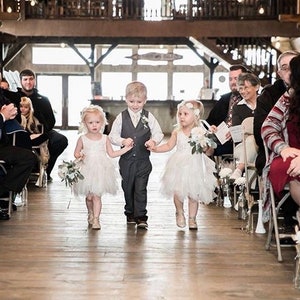 May include: Three children, two girls and a boy, walk down the aisle at a wedding ceremony. The girls are wearing white dresses and carrying bouquets of flowers. The boy is wearing a gray suit and a white vest. The children are smiling and looking at the camera.