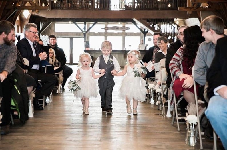May include: Three children, two girls and a boy, walk down the aisle at a wedding ceremony. The girls are wearing white dresses and carrying bouquets of flowers. The boy is wearing a gray suit and a white vest. The children are smiling and looking at the camera.