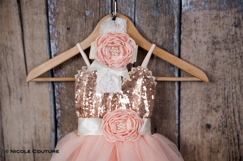 May include: A peach-colored tulle dress with a gold sequin bodice and a matching peach flower embellishment. The dress is on a wooden hanger with a white lace headband with a peach flower.
