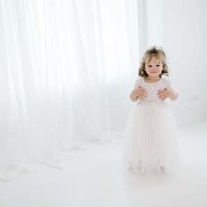 May include: A young girl in a white dress with lace details stands in a white room with white curtains. She is smiling at the camera.
