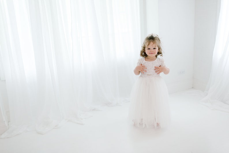 May include: A young girl in a white dress with lace details stands in a white room with white curtains. She is smiling at the camera.