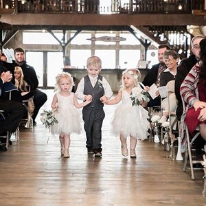 K&ouml;nnte beinhalten: Drei Kinder, zwei M&auml;dchen und ein Junge, gehen den Gang entlang bei einer Hochzeitszeremonie. Die M&auml;dchen tragen wei&szlig;e Kleider und tragen Blumenstr&auml;u&szlig;e. Der Junge tr&auml;gt einen grauen Anzug und eine wei&szlig;e Weste. Die Kinder l&auml;cheln und schauen in die Kamera.