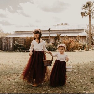 May include: Two young girls in white lace dresses and burgundy tulle skirts, holding baskets of flowers. They are standing in a grassy field in front of a wooden building.