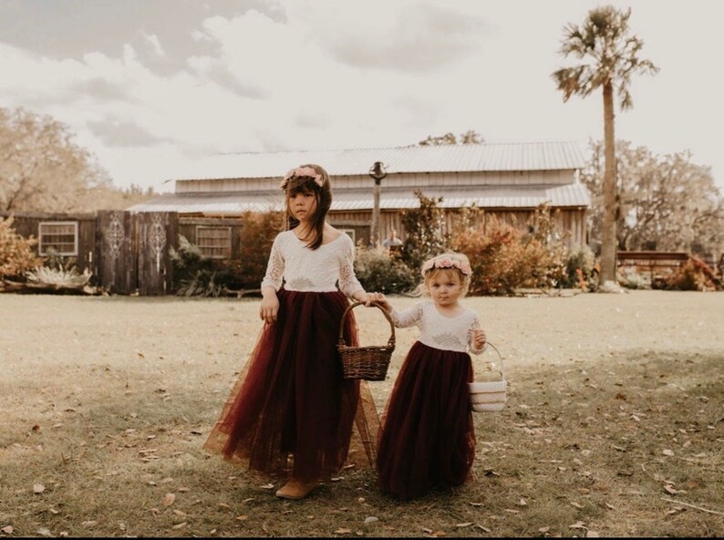 May include: Two young girls in white lace dresses and burgundy tulle skirts, holding baskets of flowers. They are standing in a grassy field in front of a wooden building.