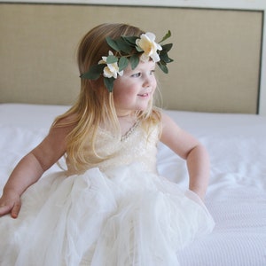 May include: A young girl wearing a white dress with a tulle skirt and a flower crown. She is sitting on a white bed and looking to the right.