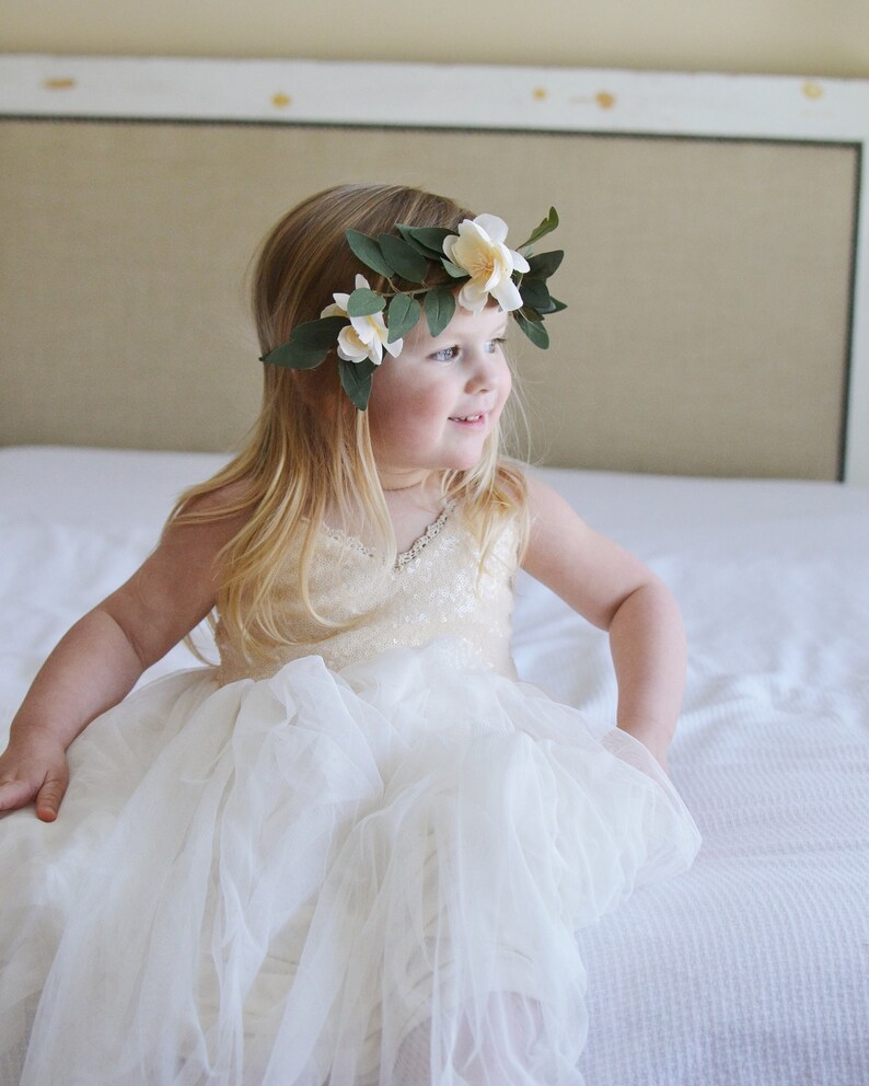 May include: A young girl wearing a white dress with a tulle skirt and a flower crown. She is sitting on a white bed and looking to the right.