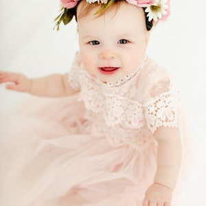 May include: A pale pink dress featuring a lace bodice with short sleeves and a layered tulle skirt. The garment is paired with a floral headband adorned with pink and white fabric flowers and green leaves, worn on the head of a young child.