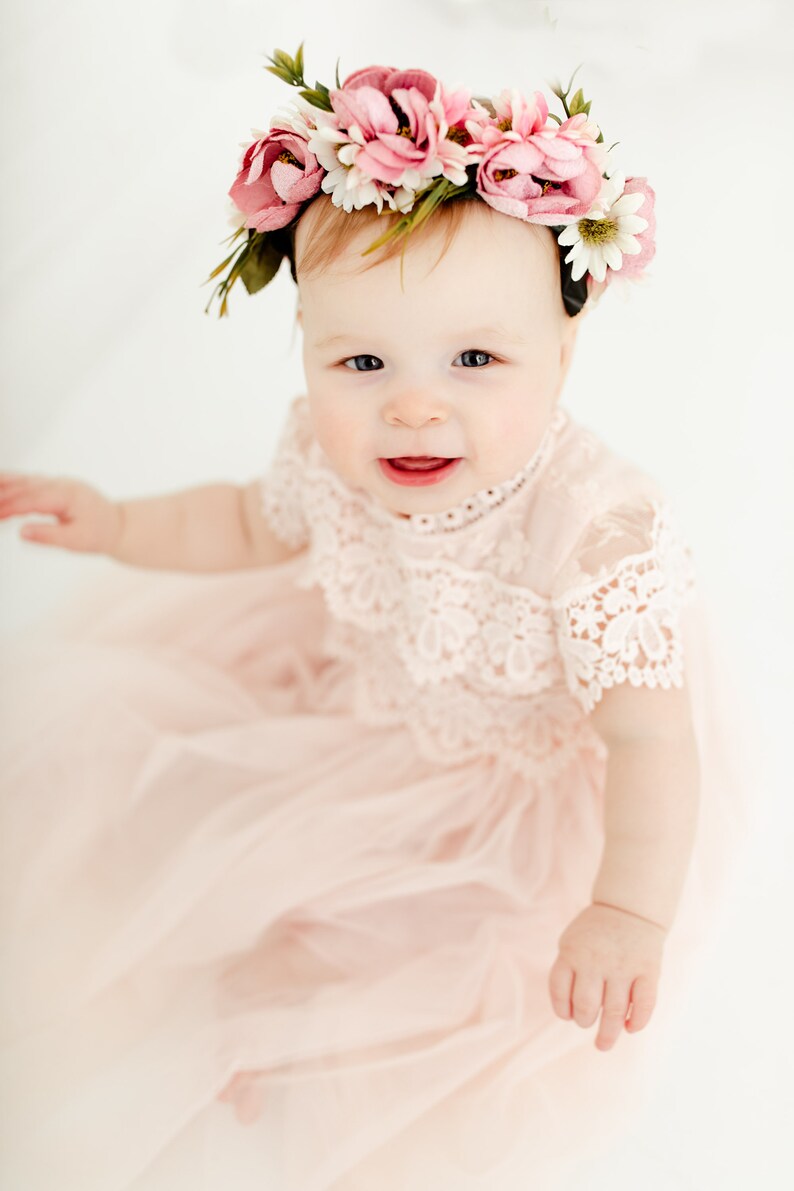 May include: A pale pink dress featuring a lace bodice with short sleeves and a layered tulle skirt. The garment is paired with a floral headband adorned with pink and white fabric flowers and green leaves, worn on the head of a young child.