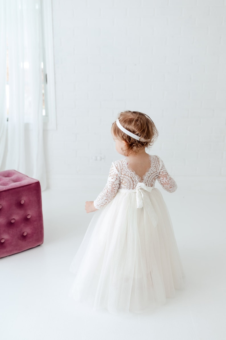 May include: A floor-length white dress with long lace sleeves and a scalloped V-neckline on the back. The bodice features intricate floral lace detailing, while the skirt is made of layered tulle. A white ribbon is tied in a bow at the waist.