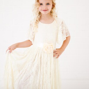 Peut inclure: Une jeune fille portant une robe en dentelle blanche avec un accent floral sur la ceinture. La robe a un col rond et des manches courtes.