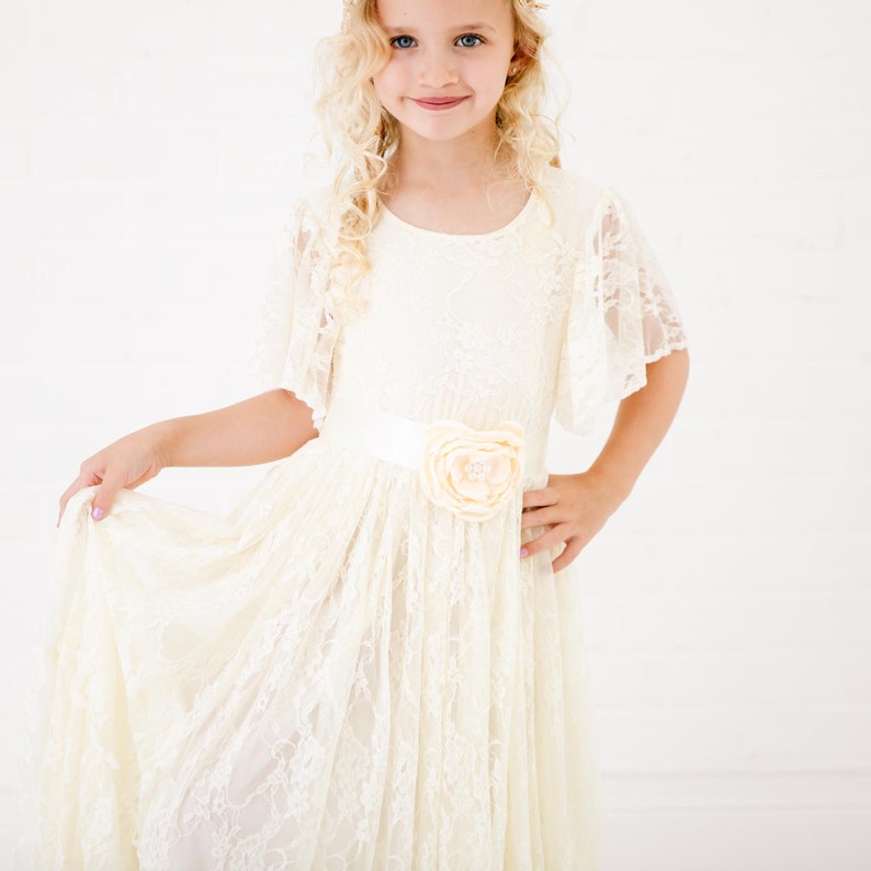 Peut inclure: Une jeune fille portant une robe en dentelle blanche avec un accent floral sur la ceinture. La robe a un col rond et des manches courtes.