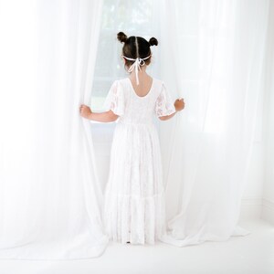 May include: A young girl in a white lace dress stands by a window, holding back the white curtains. Her hair is pulled back in two pigtails and she is wearing a white headband.