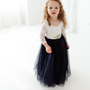 May include: A young girl wearing a white lace top with a jeweled belt and a navy blue tulle skirt.