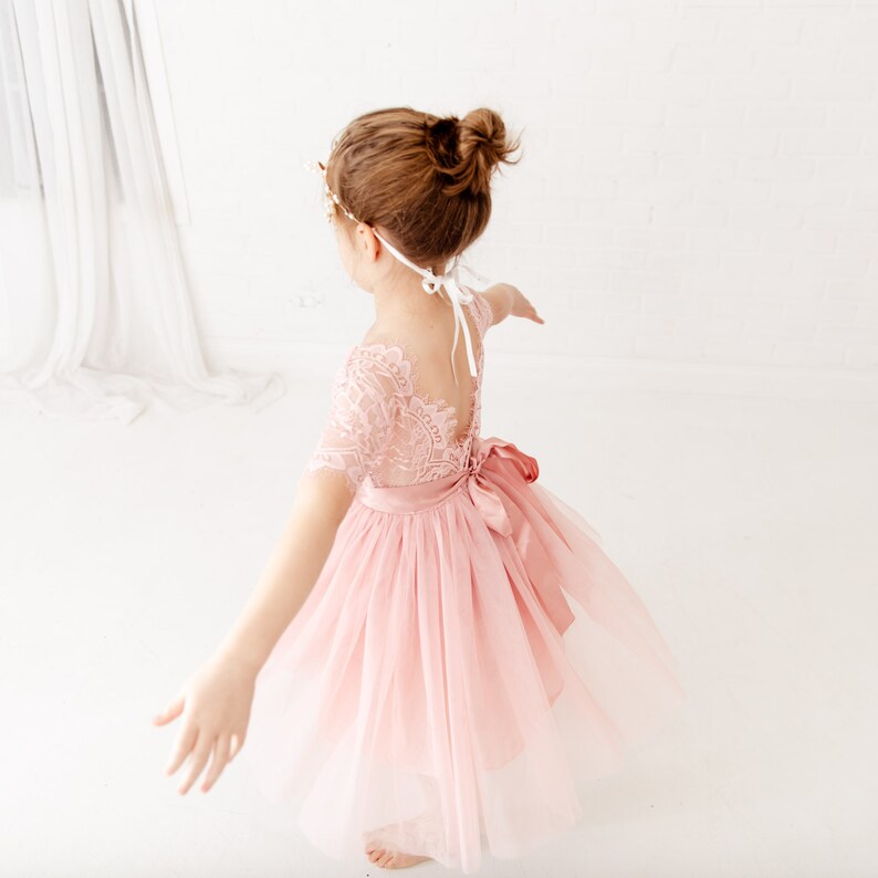 Pode incluir: Uma menina pequena vestindo um vestido de tule rosa com um corpete de renda e uma faixa de cetim branca. Ela est&aacute; usando uma faixa de cabe&ccedil;a dourada e tem o cabelo puxado para tr&aacute;s em um coque.