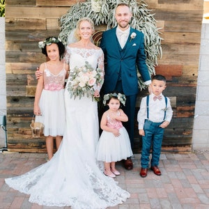 May include: A wedding ceremony featuring a bride in a white lace gown, a groom in a blue suit, and two flower girls in white dresses with pink sashes. The ceremony is taking place in front of a rustic wooden backdrop with a wreath of greenery.