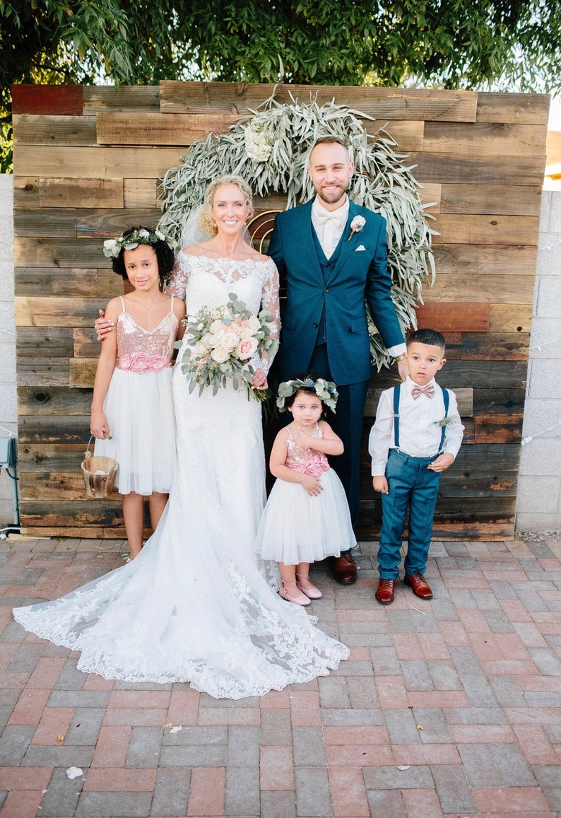 May include: A wedding ceremony featuring a bride in a white lace gown, a groom in a blue suit, and two flower girls in white dresses with pink sashes. The ceremony is taking place in front of a rustic wooden backdrop with a wreath of greenery.