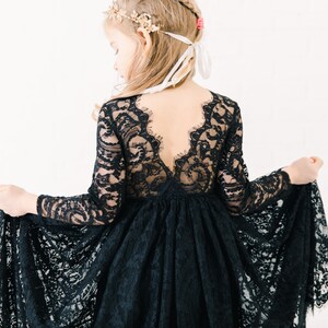 May include: A long-sleeved black lace dress featuring a deep V-shaped back neckline with scalloped edges. The bodice and sleeves are crafted from intricate floral lace, while the skirt consists of a full, flowing layer of matching black lace.