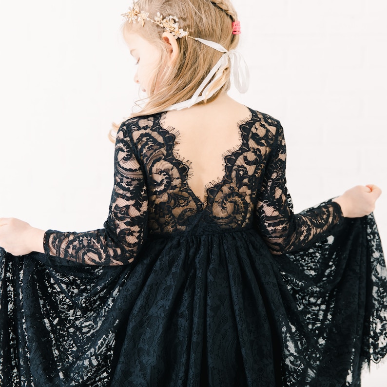 May include: A long-sleeved black lace dress featuring a deep V-shaped back neckline with scalloped edges. The bodice and sleeves are crafted from intricate floral lace, while the skirt consists of a full, flowing layer of matching black lace.