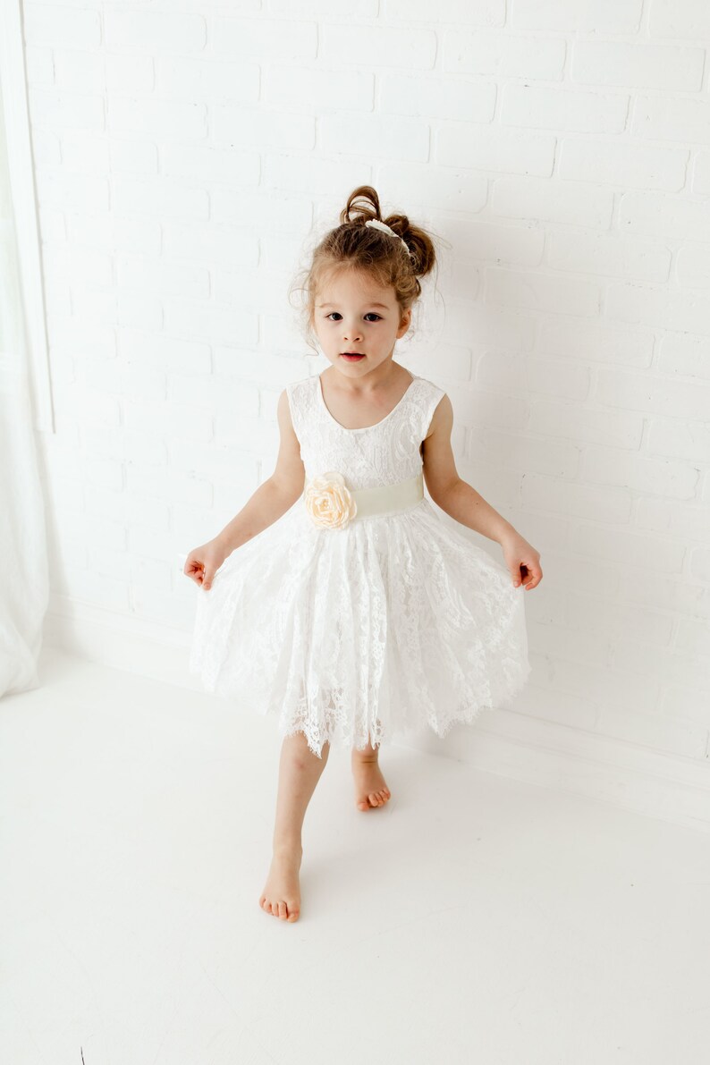 Pode incluir: Uma menina pequena vestindo um vestido branco de renda com uma faixa de cetim e um detalhe floral. O vestido tem um decote redondo e &eacute; sem mangas. A menina est&aacute; em p&eacute; em um fundo branco.