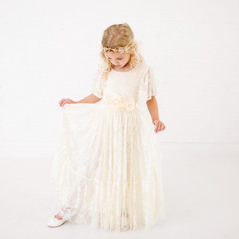 以下が含まれることがあります： 花冠と花柄のサッシュベルトをつけた白いレースのドレスを着た少女。ドレスはスカートがふっくらしていて、袖は短い。