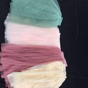 May include: Four tulle skirts in different colors: green, pink, burgundy, and beige. The skirts are layered on top of each other, with the green skirt on top and the beige skirt on the bottom.