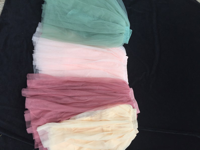 May include: Four tulle skirts in different colors: green, pink, burgundy, and beige. The skirts are layered on top of each other, with the green skirt on top and the beige skirt on the bottom.