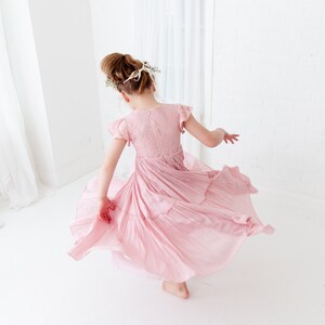 May include: A young girl wearing a pink, tiered, lace dress with a floral crown. She is twirling and her dress is flowing around her.