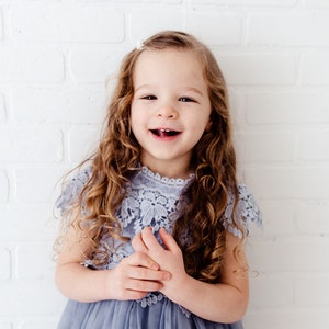 K&ouml;nnte beinhalten: Ein junges M&auml;dchen mit langen, lockigen braunen Haaren tr&auml;gt ein hellblaues T&uuml;llkleid mit einem Spitzenoberteil. Sie l&auml;chelt und schaut in die Kamera.