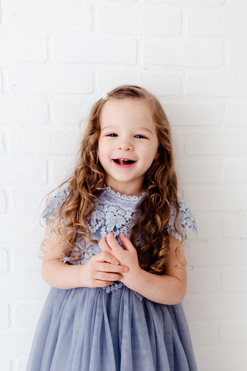 K&ouml;nnte beinhalten: Ein junges M&auml;dchen mit langen, lockigen braunen Haaren tr&auml;gt ein hellblaues T&uuml;llkleid mit einem Spitzenoberteil. Sie l&auml;chelt und schaut in die Kamera.