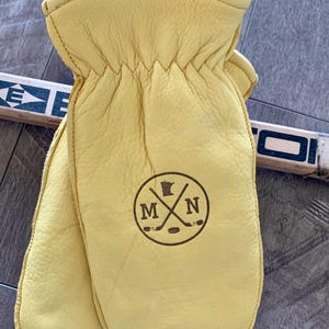 May include: A pair of yellow leather mittens with a brown leather logo featuring the outline of the state of Minnesota, two crossed hockey sticks, and the letters "M" and "N".