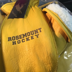 May include: A pair of yellow leather hockey gloves with a white fleece lining. The gloves have the text "ROSEMOUNT HOCKEY" embroidered on the back.