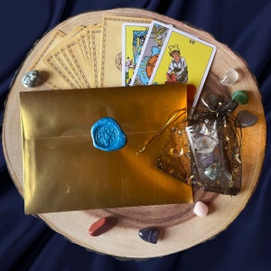 May include: A golden envelope sealed with a blue wax stamp, tarot cards, and various crystals arranged on a wooden slice. A small black mesh bag contains additional crystals. The cards feature colorful illustrations.