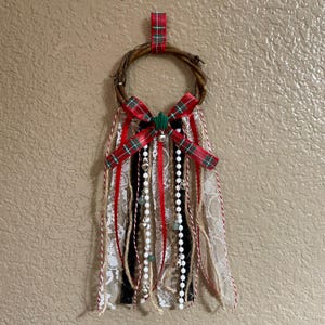 May include: A small twig wreath with a red and green tartan bow and hanging ribbons. The ribbons include red, white lace, black, and natural twine. White beaded strands and small silver bells are attached, creating a festive holiday decoration.