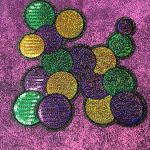 May include: A sequined applique in a dog shape, featuring green, gold, and purple circles with black borders. The applique is set against a textured purple background.