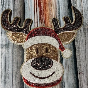 May include: A festive reindeer decoration with a Santa hat. The reindeer's face and antlers are adorned with sequins in gold, brown, and white. The Santa hat is red and white, with a red trim and a white pom-pom.
